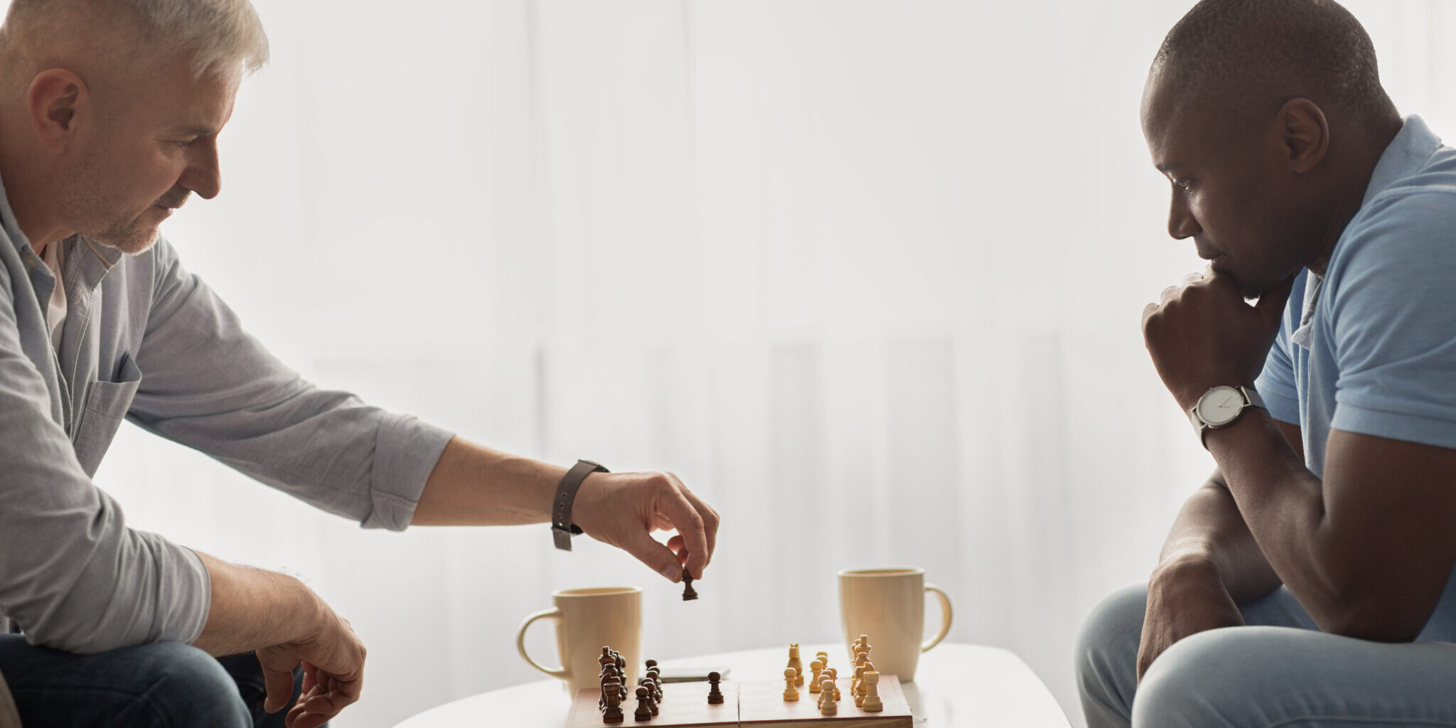 Concentrated senior man playing chess with friend, sitting at table. Pensive adult african american and caucasian males have intellectual entertainment in living room during covid-19 quarantine